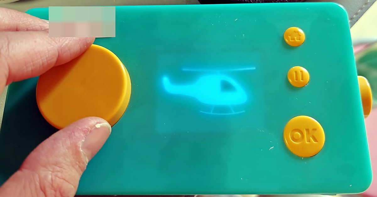 The Lunii V2 story box held in hand, its small screen glowing with a helicopter silhouette, beside the big yellow selection wheel and the home, pause and OK buttons