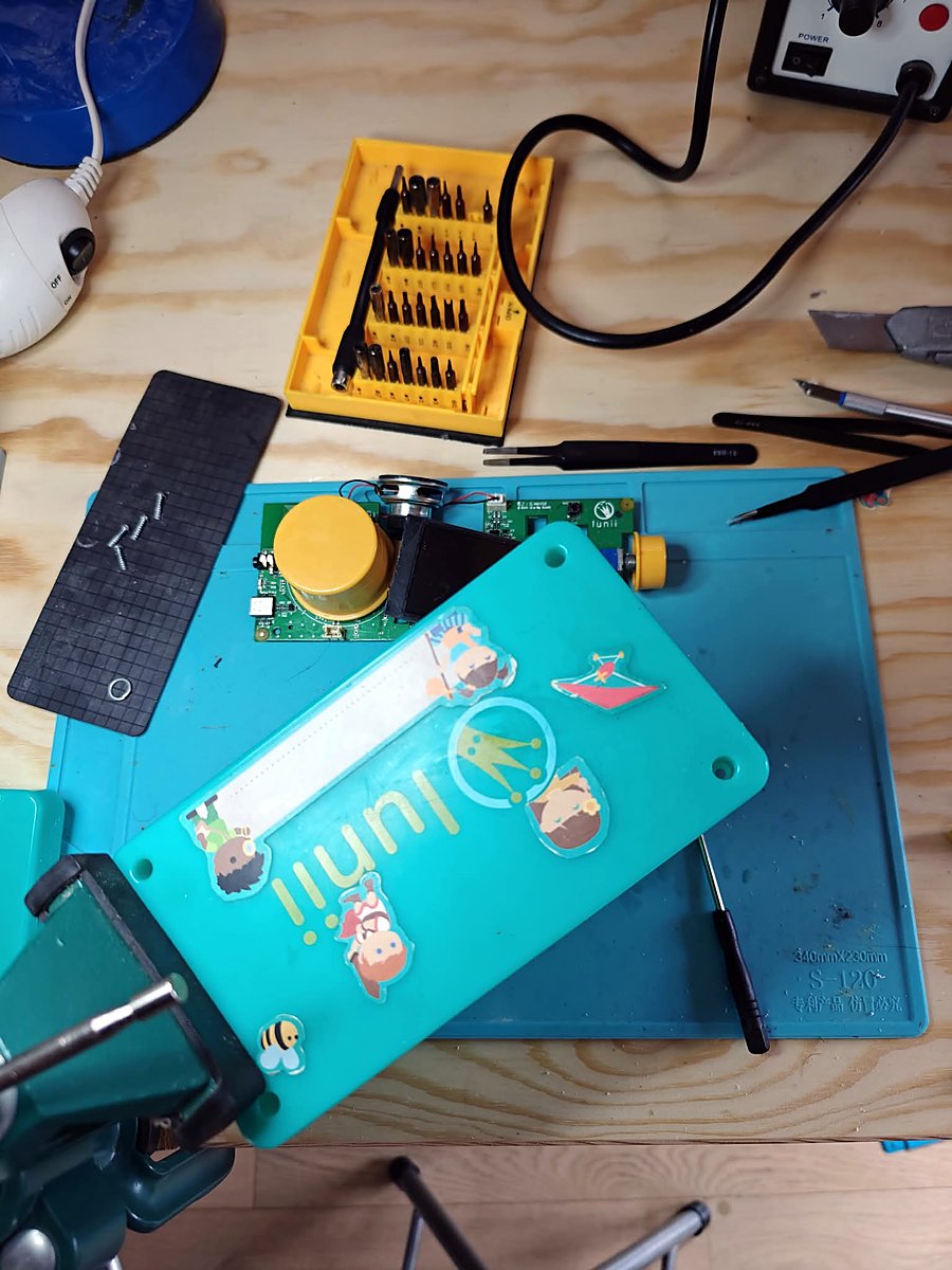 The Lunii taken apart on a blue silicone repair mat: the board with its big yellow wheel and speaker, the back shell covered in character stickers, tweezers and a bit set around it