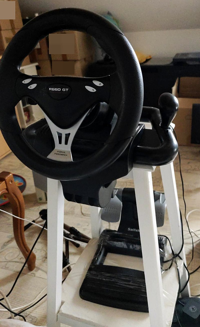 The Saitek R660 GT force feedback wheel with its gear lever, mounted on a white stool, with the pedal set on the shelf below