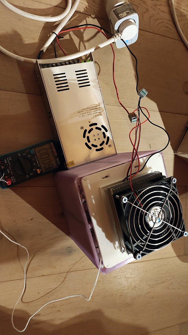 The assembled purple box with its white roof and the Peltier kit with a big fan on top, a power supply, a multimeter and a thin white wire running to the box, on a wooden floor