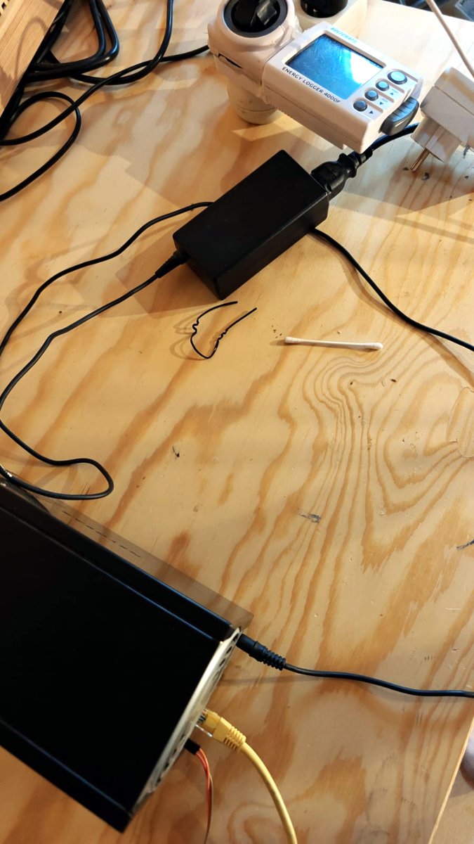 The RN102 on a wooden desk, its 12 V power brick plugged into a Voltcraft plug meter, a yellow Ethernet cable and the serial wires at the back