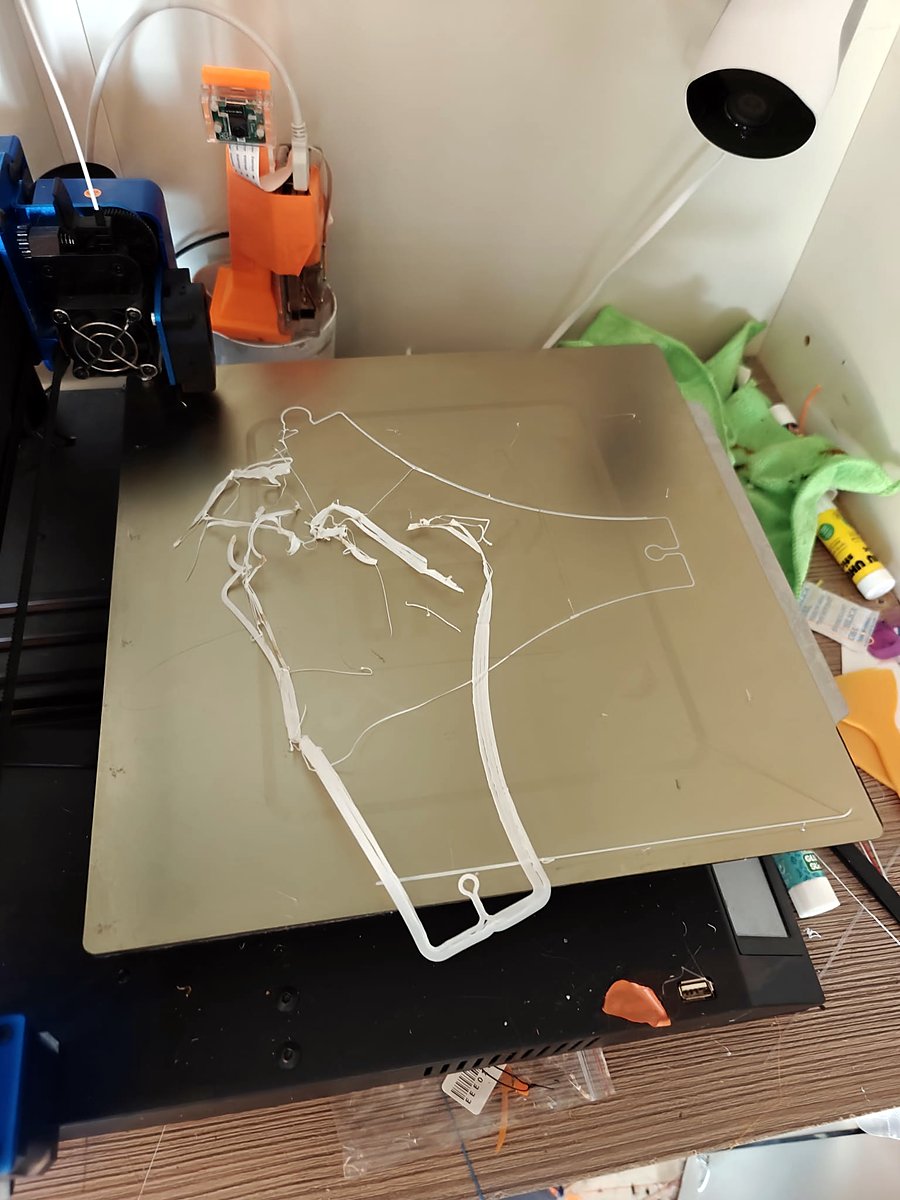 A 3D printer bed with a failed print: a thin outline of a part and many tangled white strings, with two cameras at the top