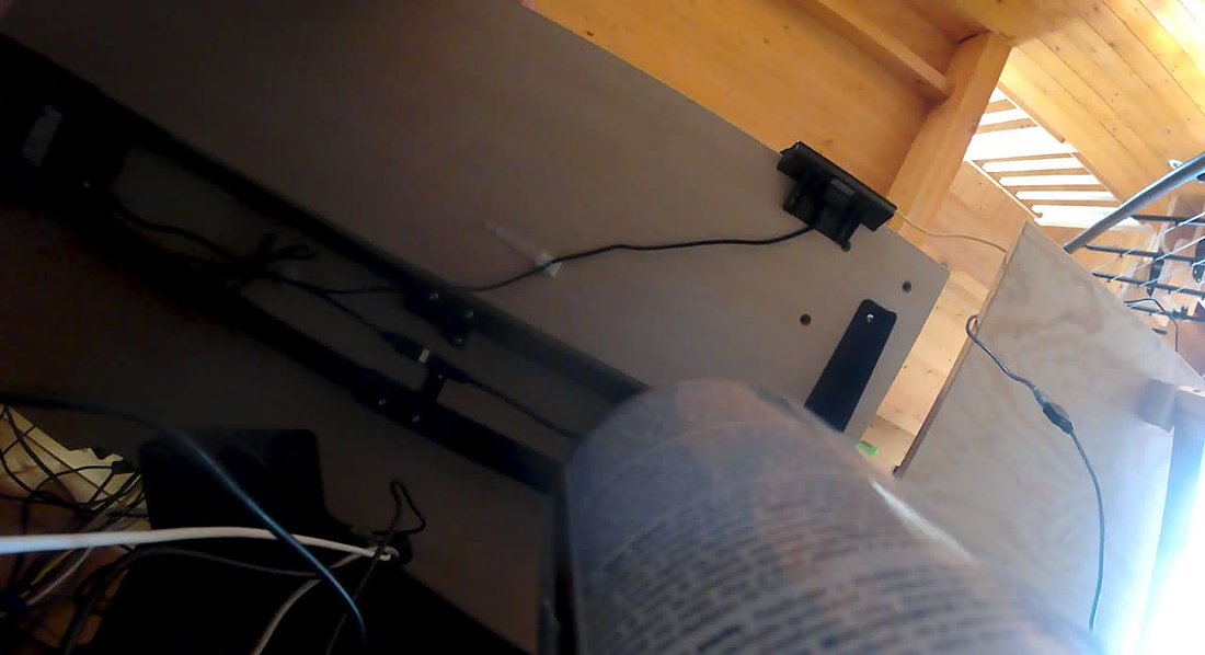 A tilted frame from the AT-03 showing the underside of a wooden desk with cables, and a book page in the foreground