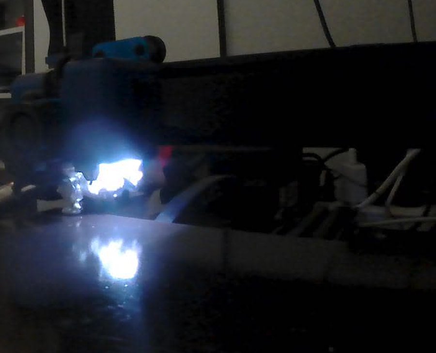 A dark frame from the AT-03 near the printer: the print head with a very bright lamp and its reflection on the bed